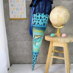 diese besondere Schultüte passend zum Ergobag Ranzen „Abtauchbär“ – farblich abgestimmt und mit einem beeindruckenden Hai-Motiv auf der Vorderseite. Perfekt für Kinder, die das Meer lieben und mit einem starken Begleiter in die Schule starten möchten. Der Name deines Kindes wird hochwertig gestickt, auf Wunsch auch mit dem Schriftzug „Schulkind 20XY“. Damit wird die Schultüte zu einem persönlichen Unikat, das perfekt zum Ranzen passt. Der obere Abschluss wird mit luftigem Tüll gefertigt und mit einem farblich passenden Satinband verschlossen – beides ist im Preis enthalten. Im Inneren sorgt ein stabiler Papprohling mit Holzspitze (70 cm Länge) für Standfestigkeit. Mit Tüllabschluss erreicht die Schultüte eine Gesamthöhe von ca. 105 cm. Nach der Einschulung als Kissen nutzbar Nach dem großen Tag kannst du den Rohling einfach entfernen. Optional biete ich dir ein Innenkissen an – so verwandelt sich die Schultüte in ein kuscheliges Erinnerungsstück, das dein Kind noch viele Jahre begleitet. Lieferumfang: Schultüte aus Stoff (70 cm Rohling, Gesamthöhe ca. 105 cm) Hai-Motiv passend zum Ergobag „Abtauchbär“ Gestickter Name (plus „Schulkind 20XY“ auf Wunsch) Tüllabschluss & Satinband zum Verschließen Papprohling mit Holzspitze Material: 97 % Baumwolle, 3 % Elastan Hinweis: Farben und Stoffzuschnitte können leicht von den Fotos abweichen – jede Schultüte ist ein handgefertigtes Unikat.